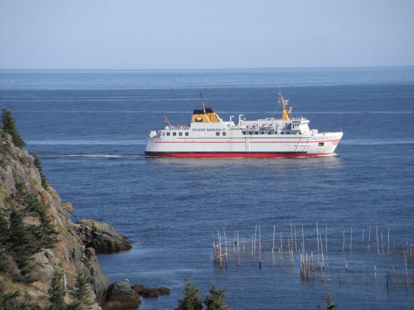 A white and red ferry is sailing in the ocean near a rocky shore.
