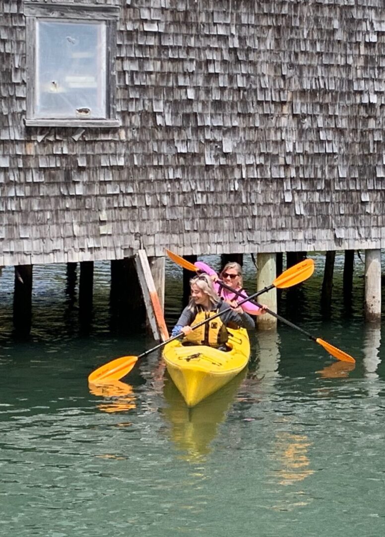 Two people paddling a yellow kayak in front of a wooden house.