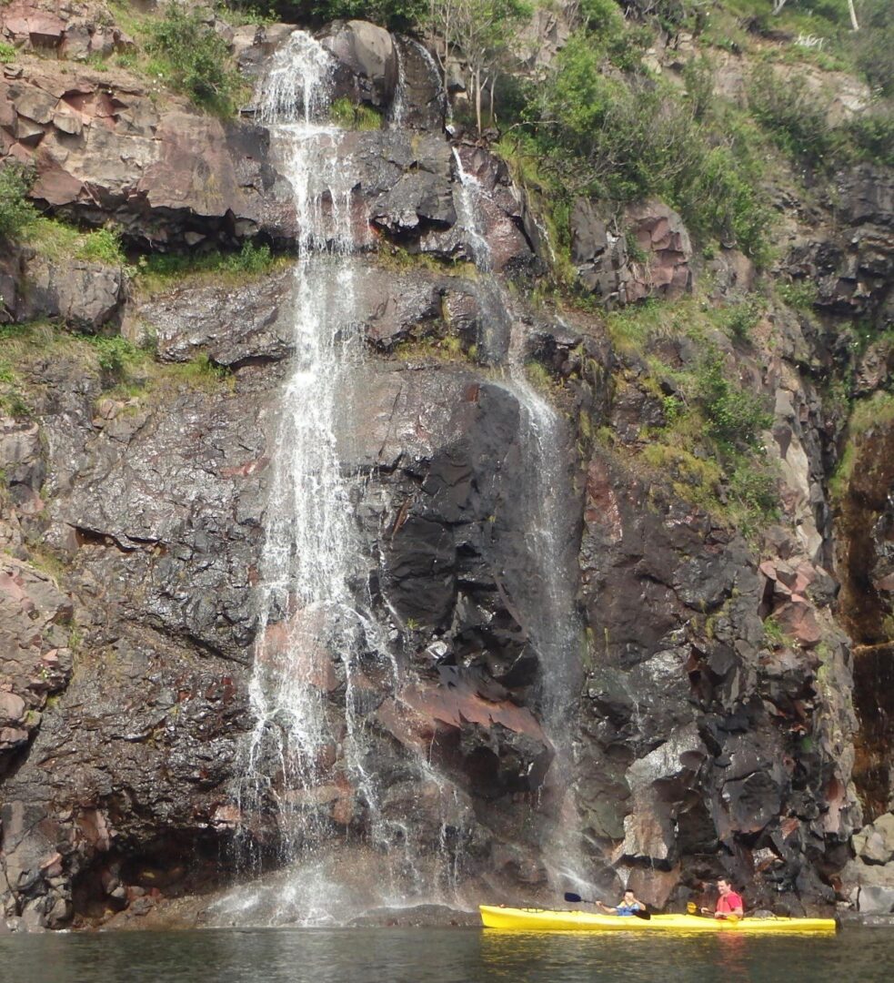 A kayaker paddles past a waterfall in front of a large rock.